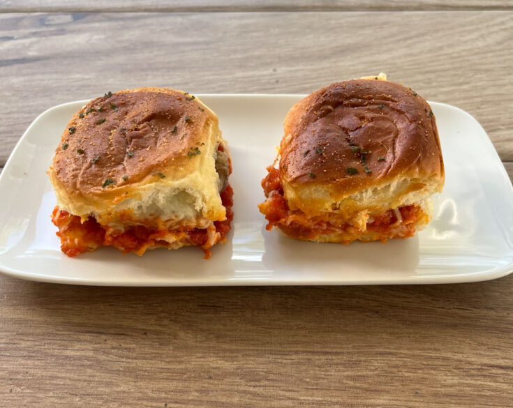 2 small rolls with a meatball, marinara sauce and cheese sitting on a white rectangular plate on top of a wood surface
