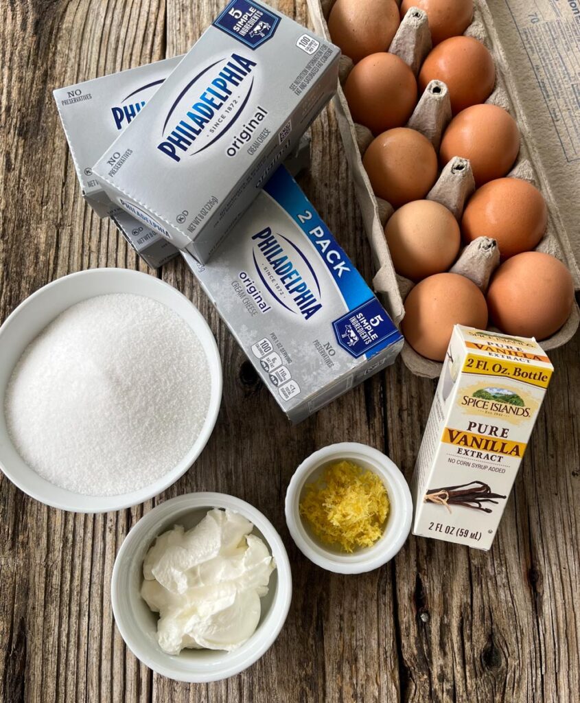 Carton of brown eggs, 4 packages of cream cheese, a box of pure vanilla extract, and 3 white bowls with sugar in one bowl, sour cream in the second bowl and lemon zest in the third. All items are sitting on top of a wooden surface.