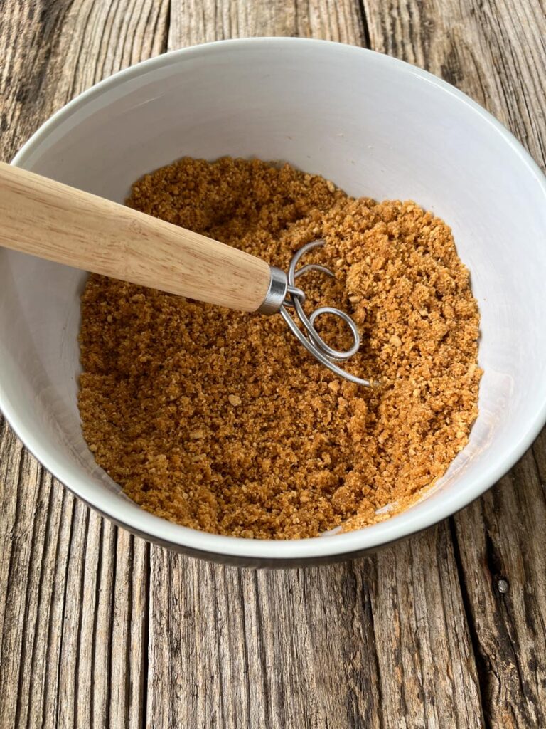 Graham cracker crumbs, sugar, and melted butter mixed together in a large white bowl with a wood handled spatula in the bowl sitting on a wooden surface.