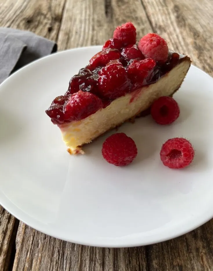 A slice of cheesecake with a raspberry sauce topping, with 3 fresh raspberries beside the slice, sitting on a white round plate. There is a gray napkin next to the plate. All sitting on a wood surface