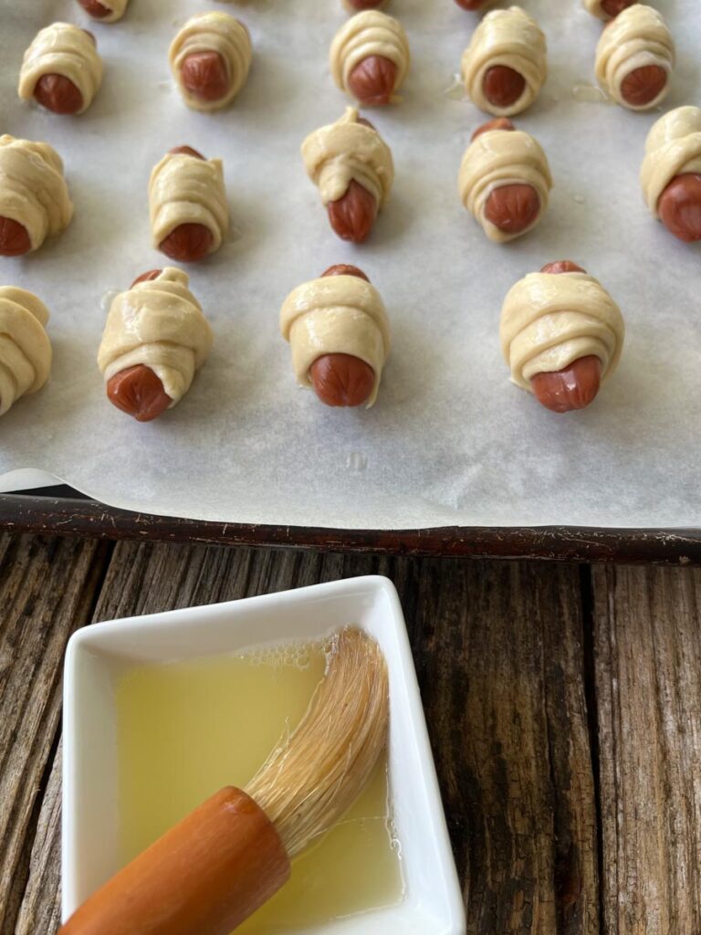 Cocktail franks rolled up in strips of crescent dough on a parchment paper lined baking pan with a small square bowl in front of pan that has melted butter and a pastry brush in it, on a wood surface