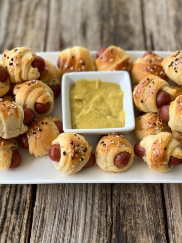Twenty baked pigs in a blanket on a white rectangular plate with a white square bowl in the center of the plate with mustard sauce in it, sitting on a wood surface