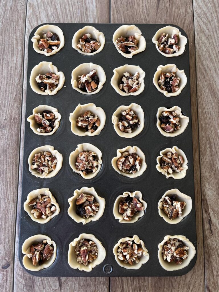 24 count mini muffin pan with uncooked pastry crust in them with chopped pecans and raisins in each cup. Pan is sitting on a wood surface