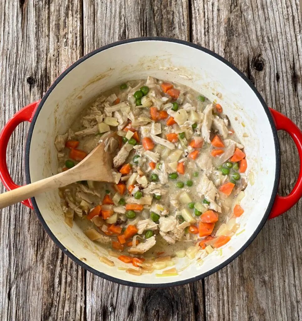 Medium dutch oven with a creamy vegetable and turkey sauce in it. The vegetables are: peas, carrots, celery, parsnip, onions, shallots, and potatoes. There is a wooden mixing spoon resting inside the dutch oven. The dutch oven is on a wood surface.