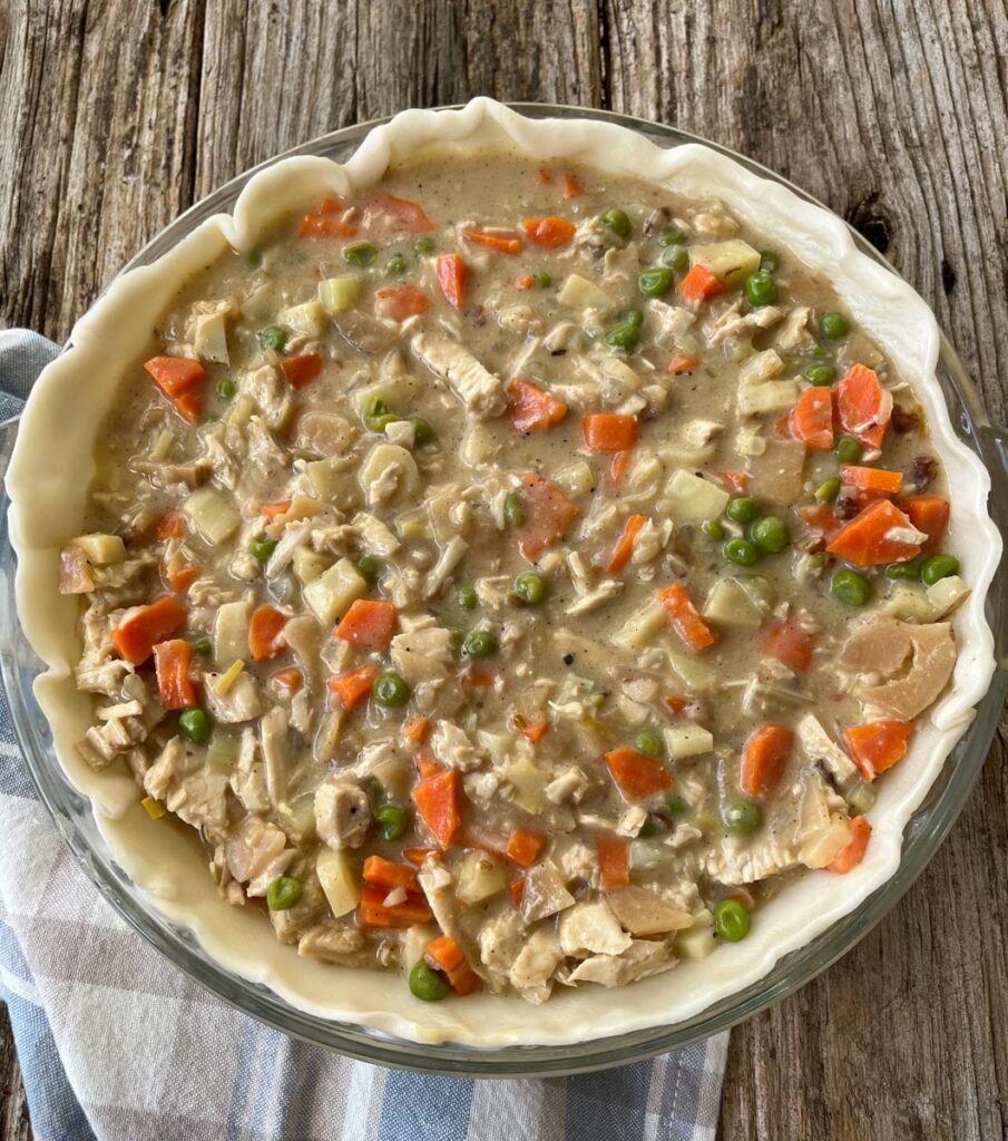Creamy vegetable and turkey filling in a deep dish pan with an uncooked pie crust. The vegetables are: peas, carrots, celery, parsnip, onions, shallots, and potatoes. The pan is sitting on a plaid linen napkin on a wood surface.