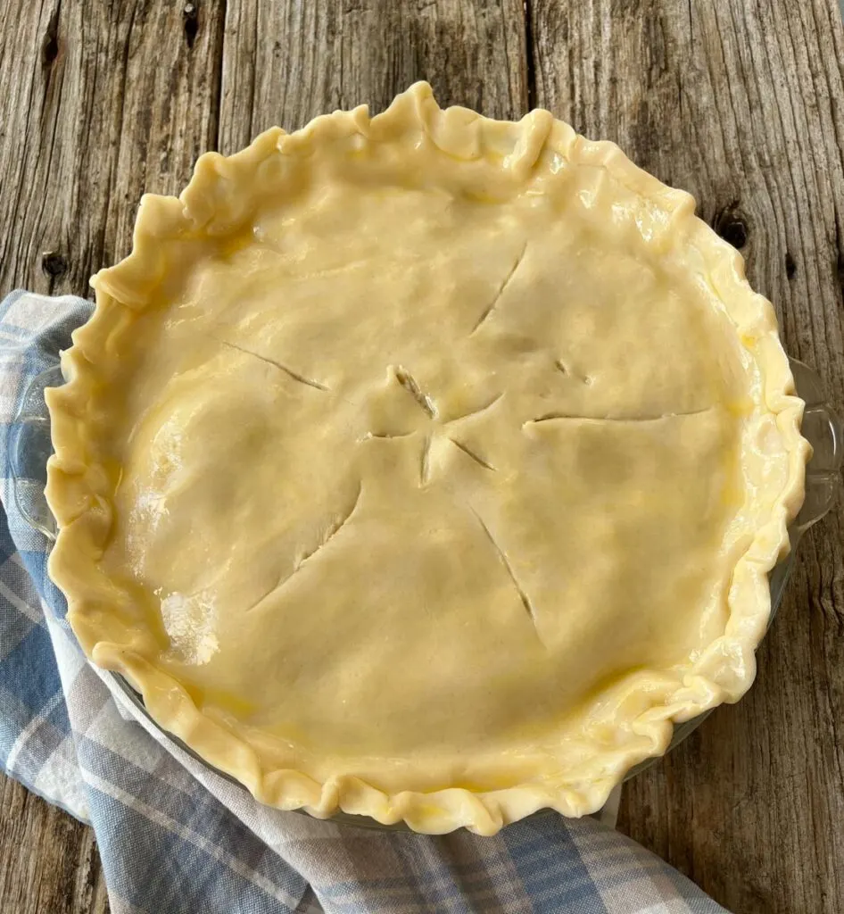 Unbaked turkey pot pie with a top crust. The top crust has an egg wash coating and thin slits in it. The pan is sitting on a plaid linen napkin on a wood surface.