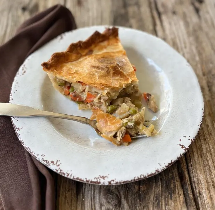 Serving of turkey pot pie on a small white round plate. There is a fork on the plate. There is a brown cloth napkin on the side of the plate. Items are on a wood surface.