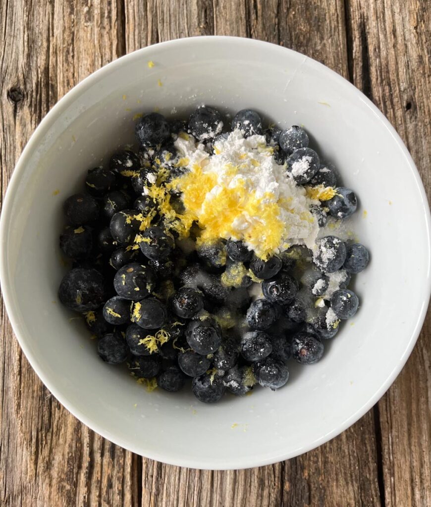 White mixing bowl with fresh blueberries in it. There is white sugar, cornstarch, lemon juice, and lemon zest poured on top of the blueberries. The bowl is sitting on a wood surface.