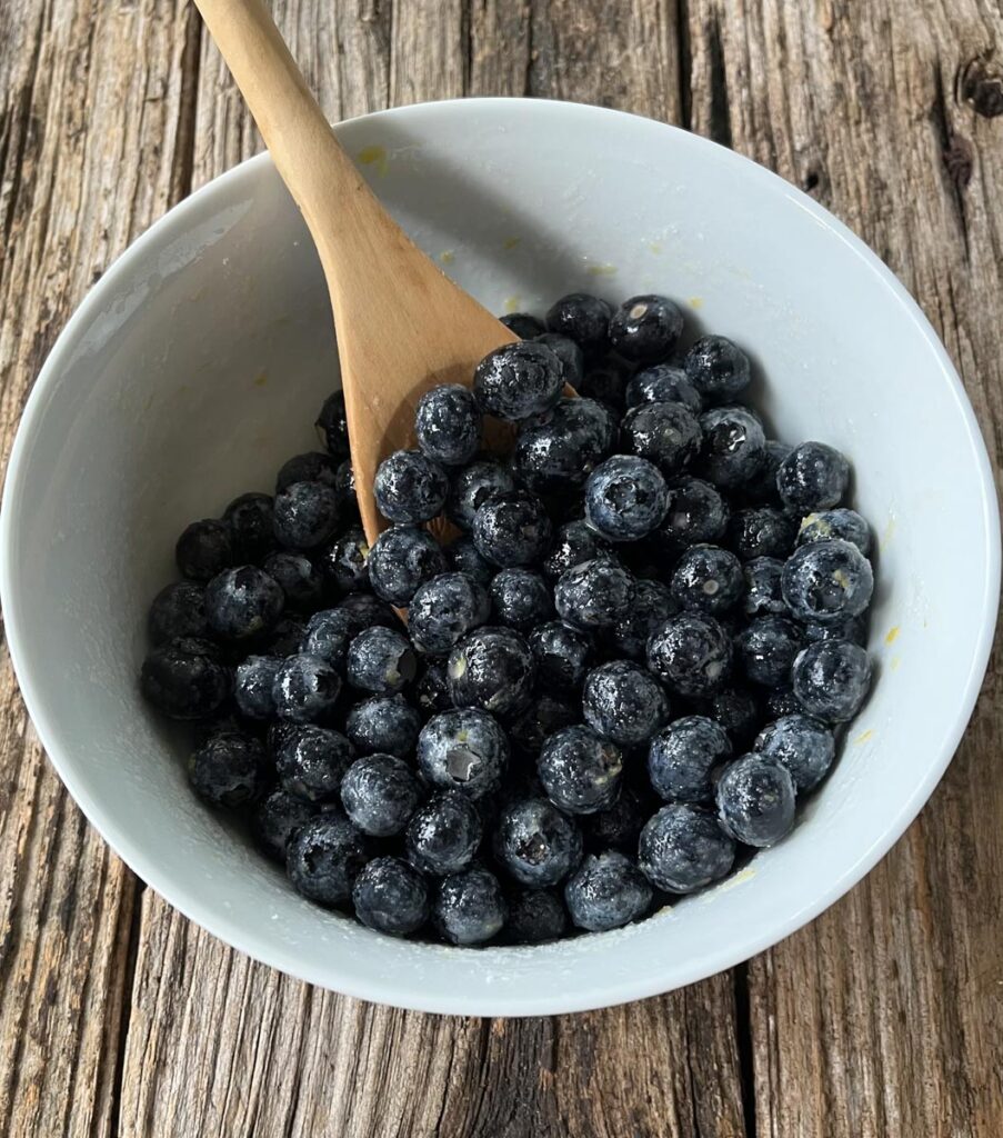 White mixing bowl with fresh blueberries coated with sugar, cornstarch, lemon juice, and lemon zest. There is a wooden spoon leaning inside the bowl. The bowl is sitting on top of a wood surface.