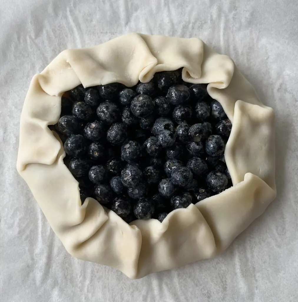 Sugar coated blueberries in the center of a rolled out pie crust. The edges of pie dough are folded up towards the center and crimped every 2 inches. It is on parchment paper.