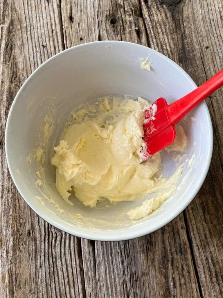 Large white mixing bowl with creamed butter in it. There is a red rubber spatula in the bowl resting on the side. Bowl is on a wood surface.