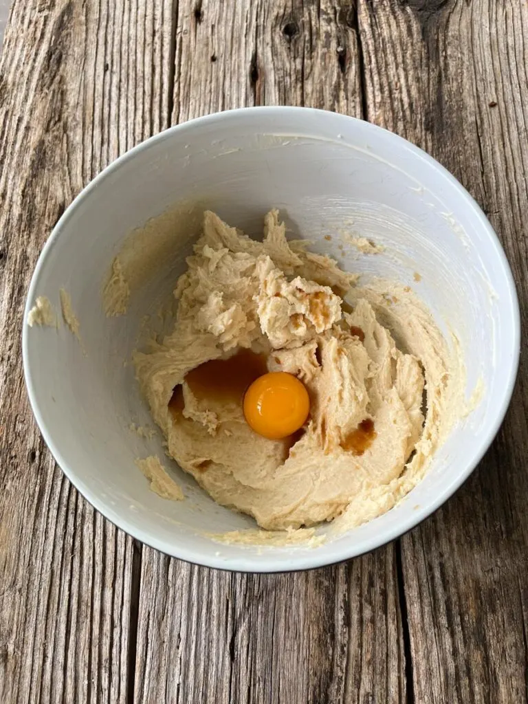 Large white mixing bowl with butter , white sugar, and light brown sugar blended together. There is an egg yolk and vanilla extract on top of the mixture. The bowl is on a wood surface.