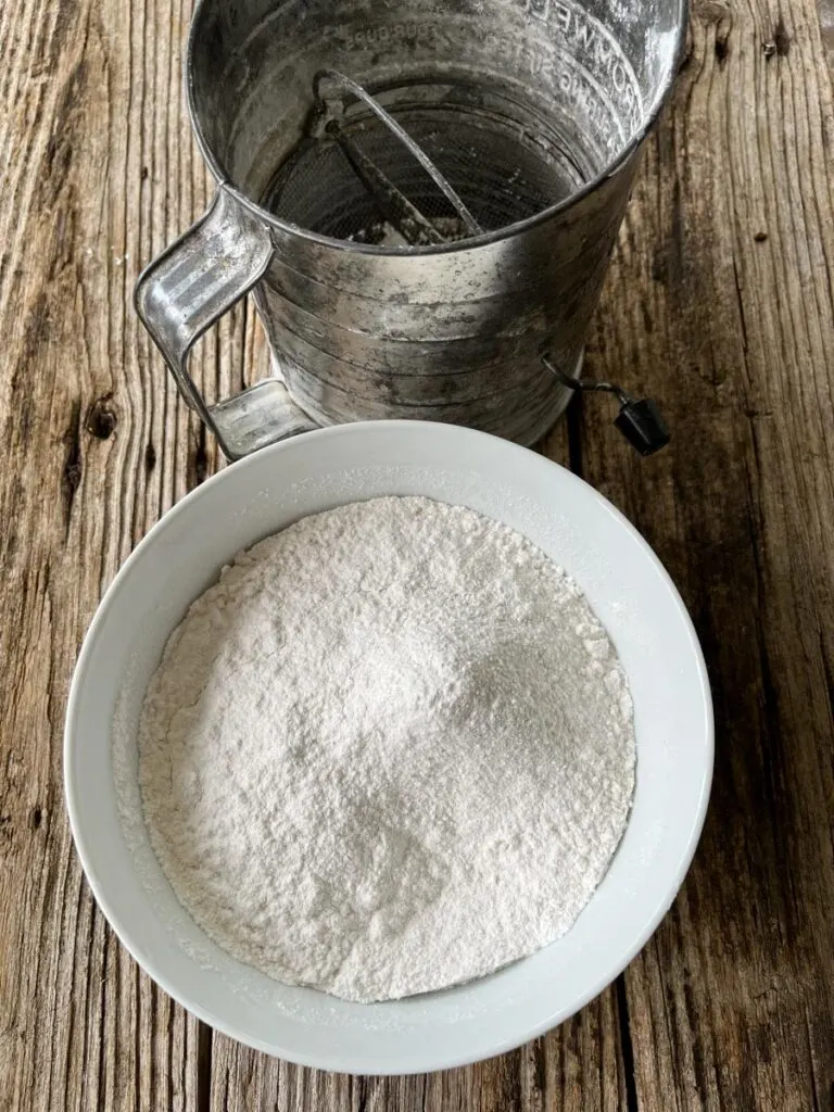 Medium size white mixing bowl with sifted flour and cornstarch in it. There is a metal sifter in the back ground. Items are on a wood surface.