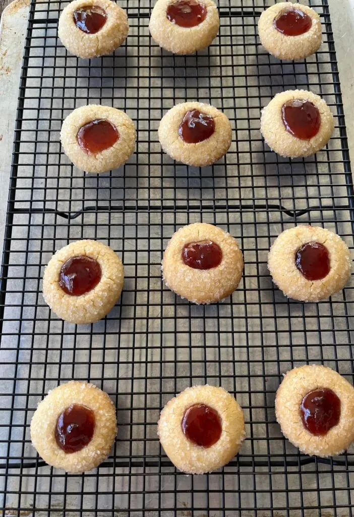 There is a dozen of sugar coated baked cookies with raspberry jam in the center of each ball on a black wire cooling rack. The rack is on a baking sheet.