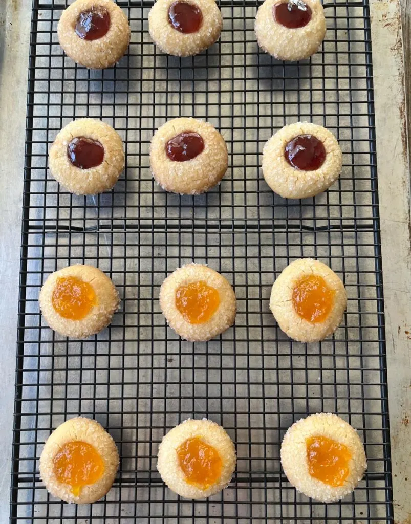 There is a dozen of sugar coated baked cookies on a black wire cooling rack. Six of the cookies have mango jam in them and the other six have raspberry jam in them. The rack is on a baking sheet.