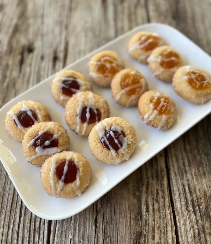 One dozen of round sugar cookies on a white rectangular plate. Six of the cookies have raspberry jam in the center of them. The other six cookies have mango jam in the center of them. All of the cookies have a white glaze drizzled on them. The plate is on a wood surface.