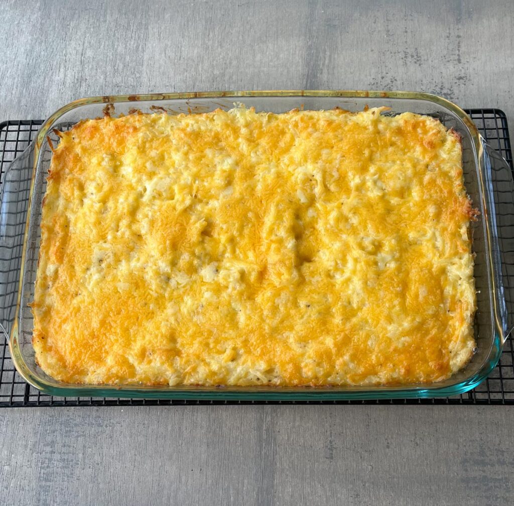 A 9" x 13" glass dish with baked hash brown casserole in it, with melted cheese on the top. The baking dish is on a black wire cooling rack sitting on a wood surface.