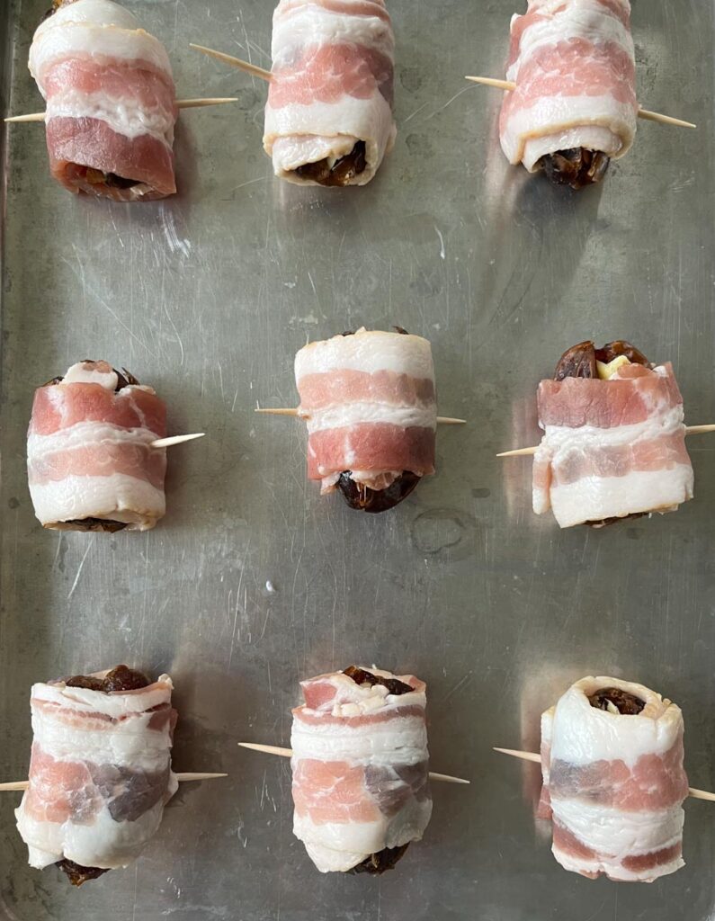 There are 9 bacon-wrapped dates stuffed with almonds on a baking sheet. Each wrapped date have a toothpick speared through it.