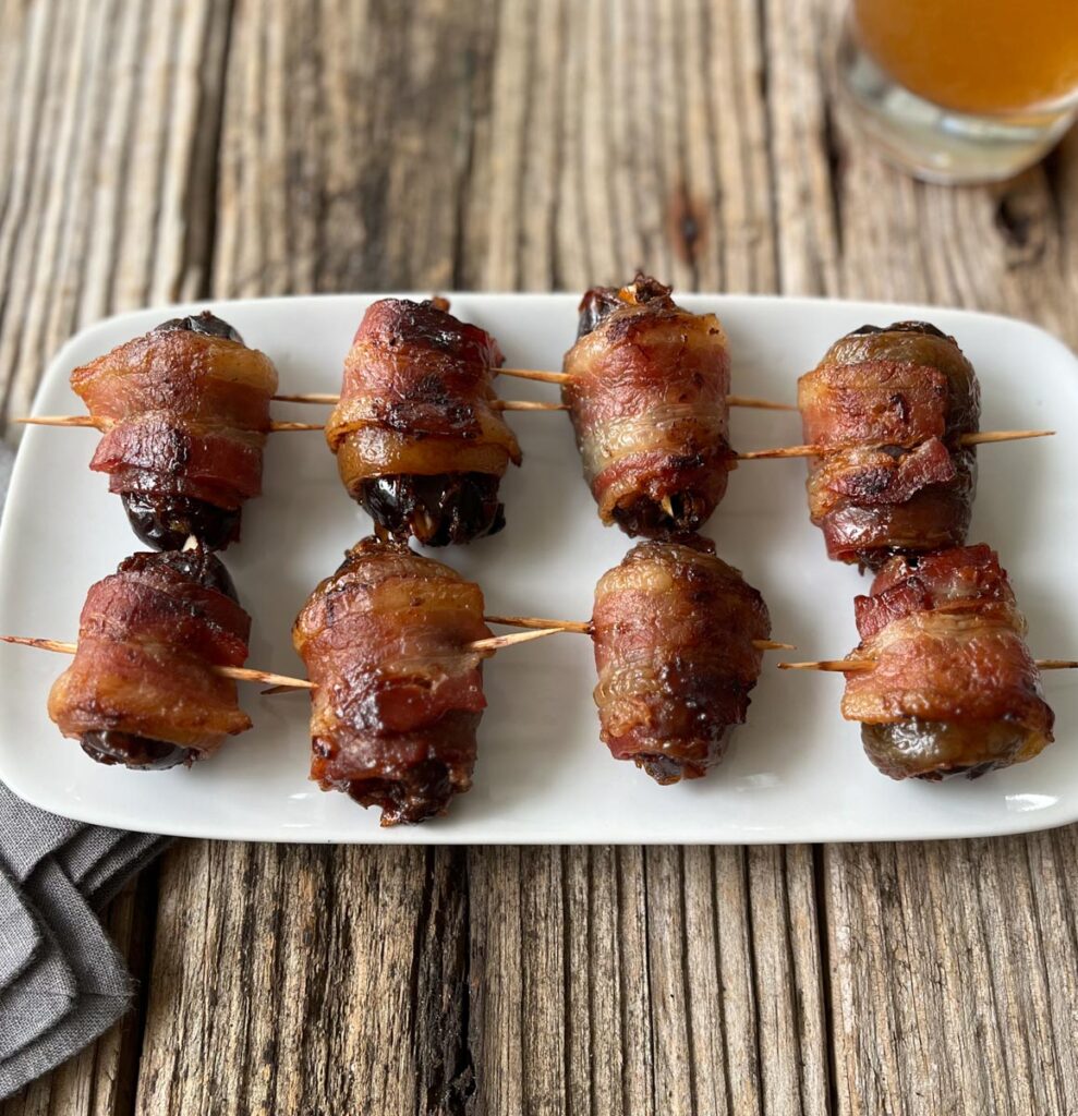 8 cooked bacon-wrapped dates stuffed with almonds arranged on a white rectangular plate. There is a glass of beer in the background and a gray cloth napkin by the side of the plate.Plate is on a wood surface.