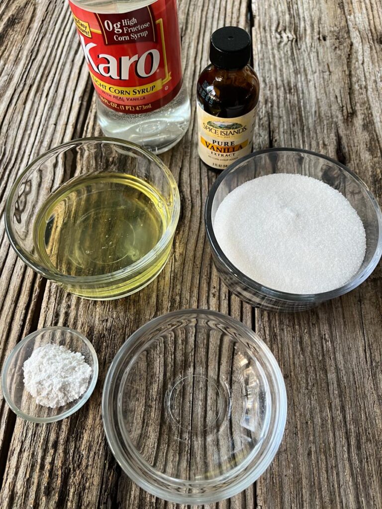 3 small glass bowls. One contains sugar, one contains egg whites, the third has water in it. There is 1 small clear glass ramekin with cream of tartar in it. There is a bottle of pure vanilla extract and a bottle of corn syrup in the background. Items are on a wood surface.