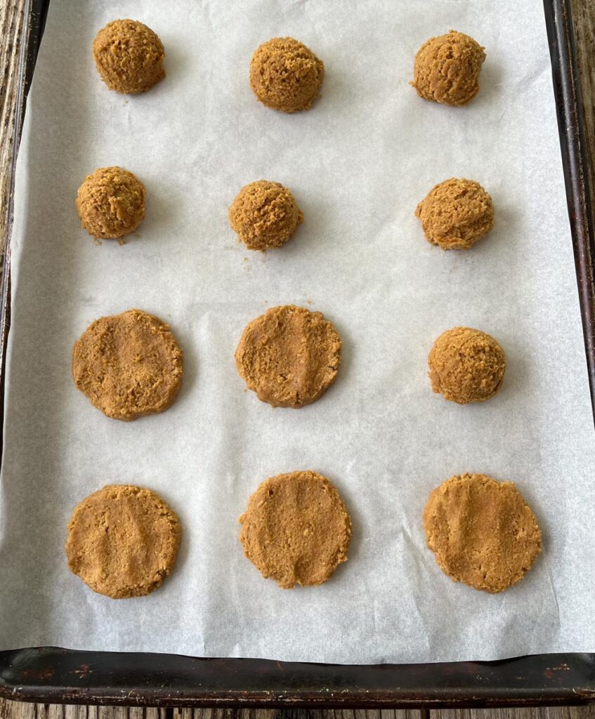 12 cookie dough balls on a parchment lined baking pan. 6 of the dough balls are slightly flattened. Baking pan is on a wood surface.