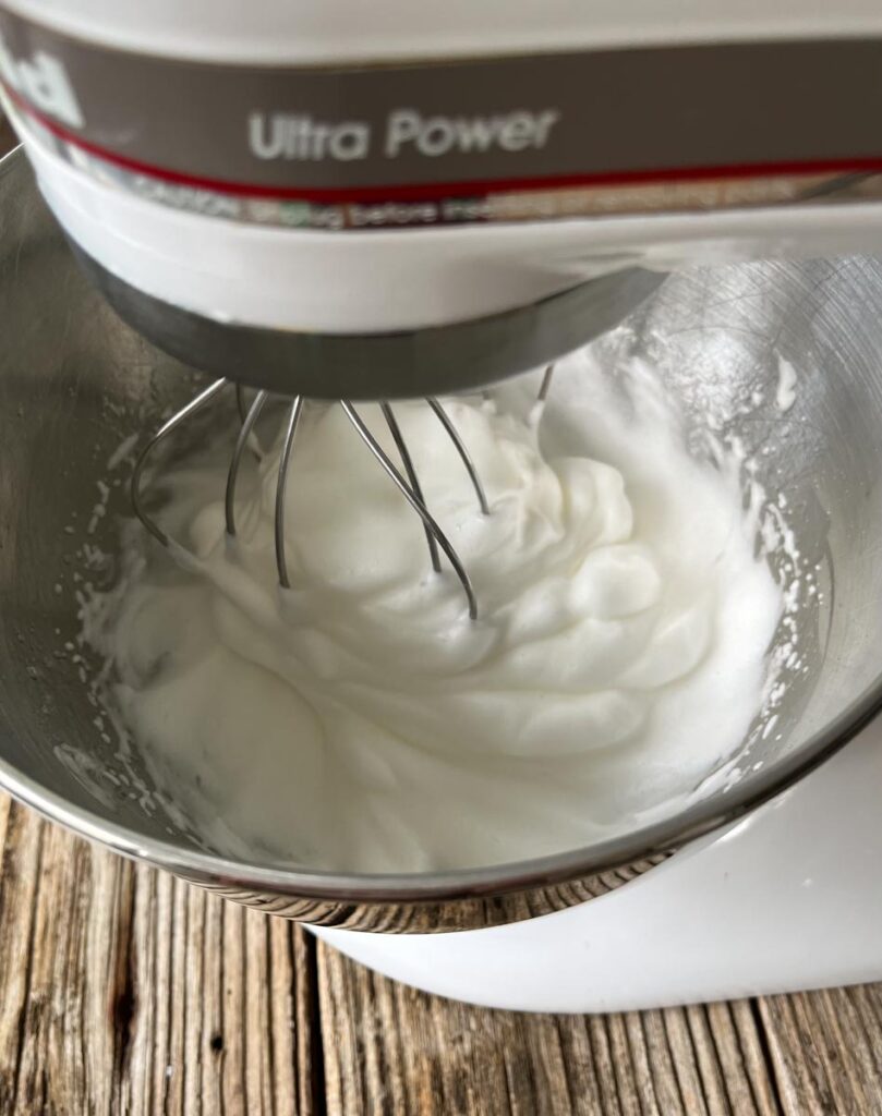 Standing mixer with whipped egg whites in the mixer bowl. Mixer is on a wood surface.