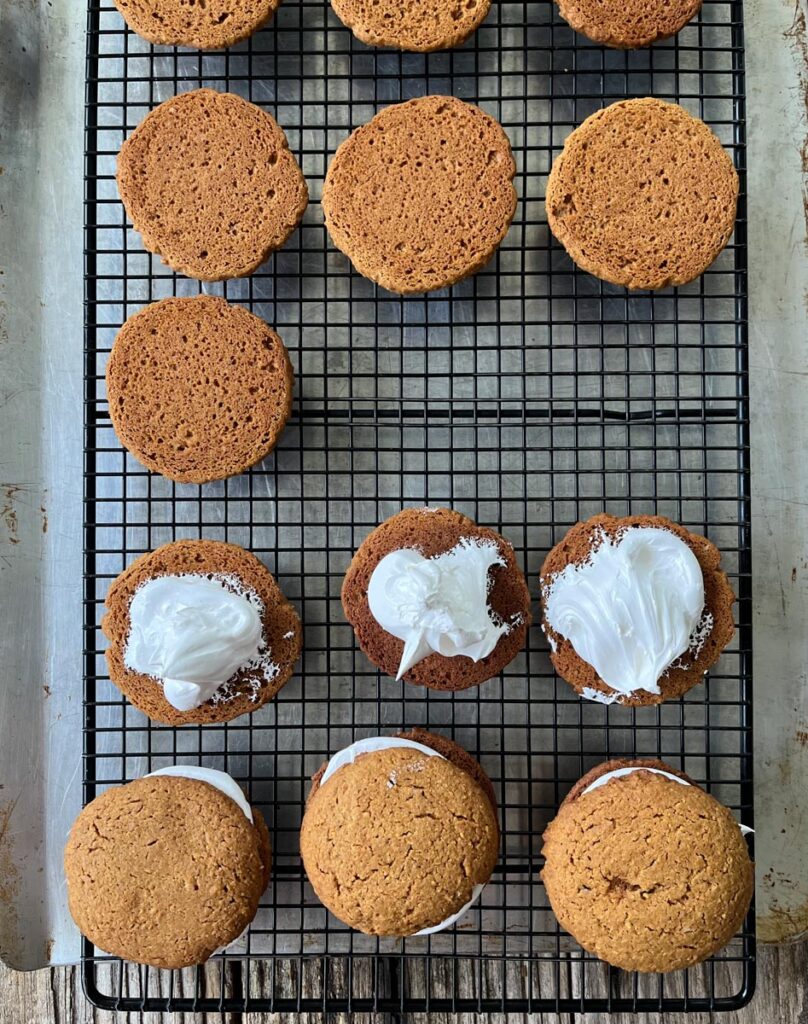 Cookies on a black wire rack. 3 cookies are assembled as sandwiches, 3 cookies have marshmallow filling on them. The remaining 6 cookies are turned upside down. Bottoms are facing up. Wire rack is on a cookie sheet. Cookie sheet is on a wood surface.