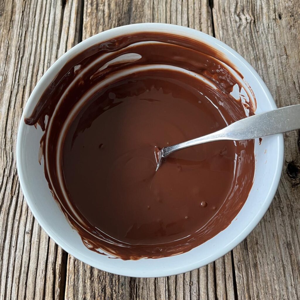 Medium size white mixing bowl with melted chocolate in it. There is a stainless spoon resting in the bowl. Bowl is on a wood surface.