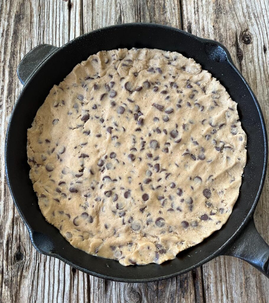 Large cast iron skillet with cookie dough pressed into the bottom. Skillet is on a wood surface.