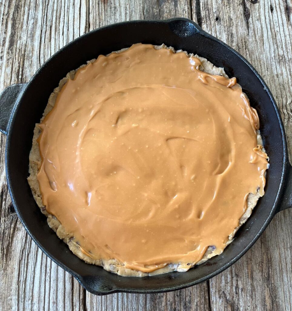 A large cast iron skillet with cookie dough on the bottom. There is a layer of melted caramel on top of the cookie dough. The skillet is on a wood surface.