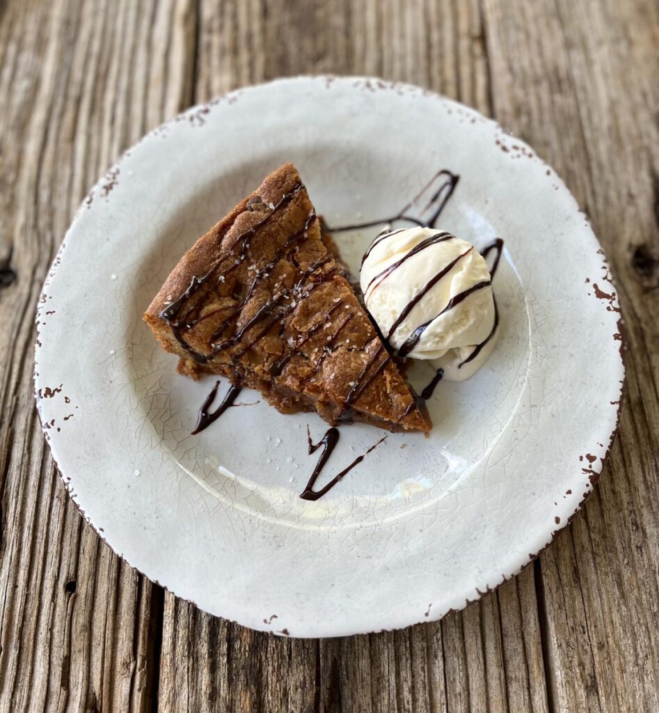 There is a white plate with a slice of Deep Dish Chocolate Chip Cookie with a scoop of vanilla ice cream and chocolate sauce drizzled over it. Plate is on a wood surface.