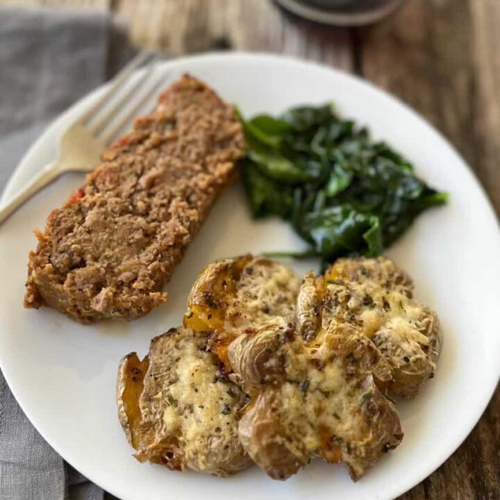Roasted smashed garlic parmesan cheese baby yellow potatoes, a slice of meatloaf, and sauteed spinach on a white round plate