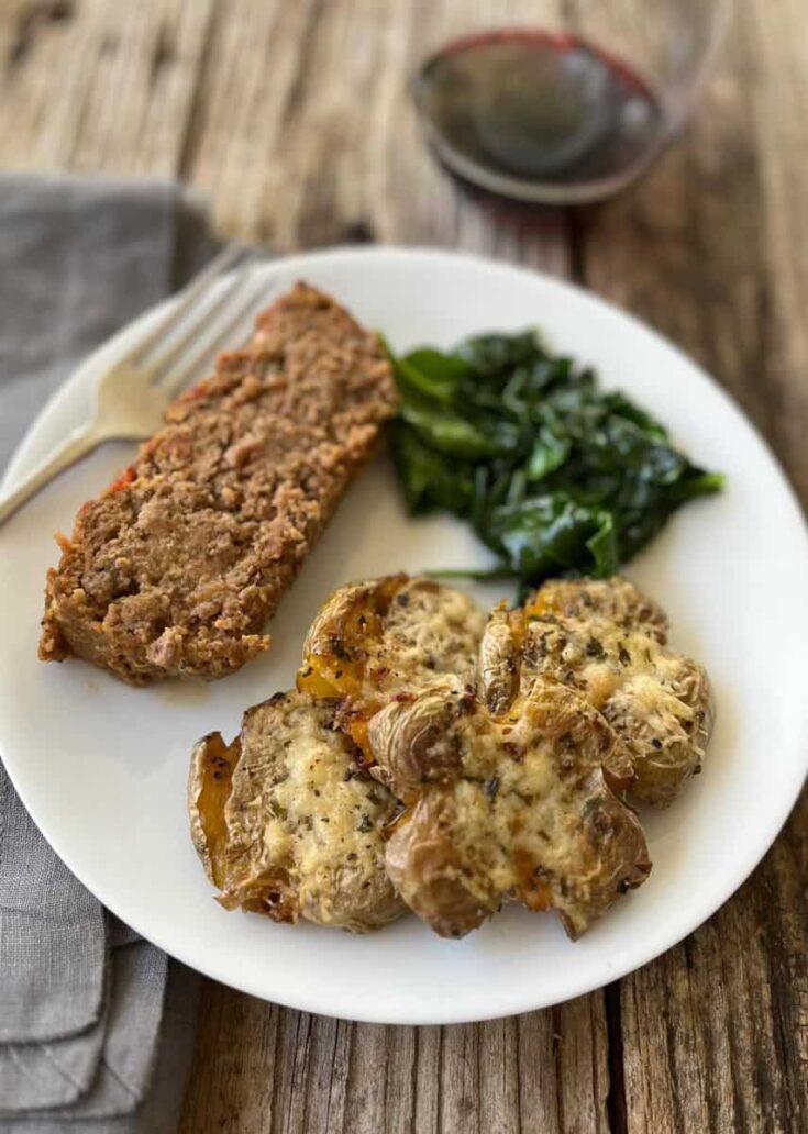 Roasted smashed garlic parmesan cheese baby yellow potatoes, a slice of meatloaf, and sauteed spinach on a white round plate