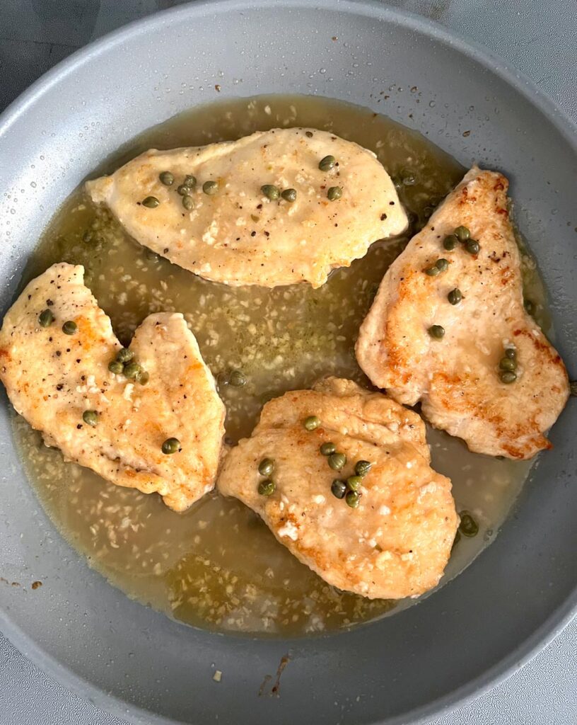Four skinless boneless chicken breasts in chicken broth, lemon juice with capers and minced garlic cooking in a large gray non-stick skillet.