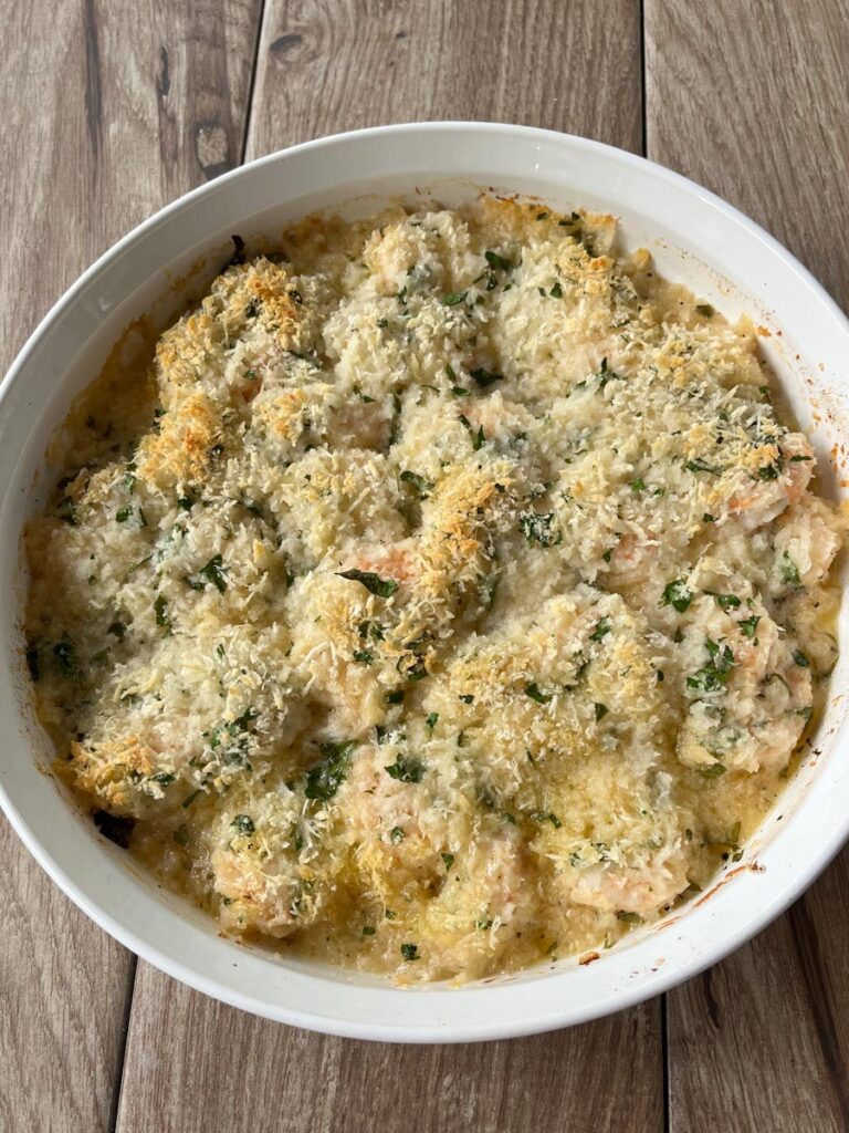 White round shallow baking dish with baked shrimp, wine, lemon juice, garlic, butter, black pepper, and hot sauce.The shrimp has a panko parmesan bread crumbs mixture sprinkled over the entire top. Dish is on a wood surface.