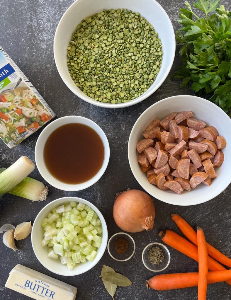 There is a bowl of dried split peas, a bowl of cut up sausage, a bowl of chopped celery and a bowl of dissolved beef bouillon. There is a box of chicken broth. There are two leeks, 2 cloves of garlic and 2 bay leaves. There are 4 carrots, stick of butter, a bunch of parsley, and an onion. There are two small ramekins with spices in them. All the items are on a gray surface.