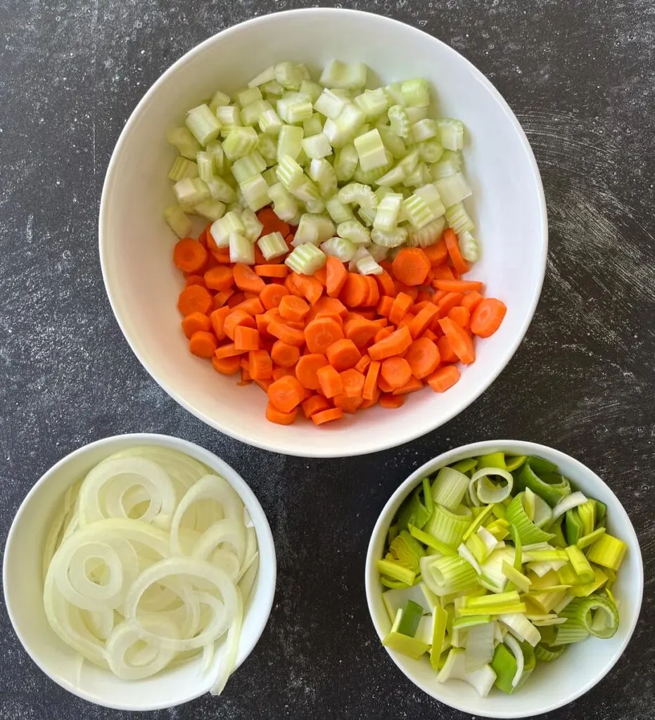 Three white bowls. Largest bow has chopped carrots and celery in it. One of the smaller bowls has sliced onion in it. The other bowl has sliced leeks in it. Bowls are on a gray surface.