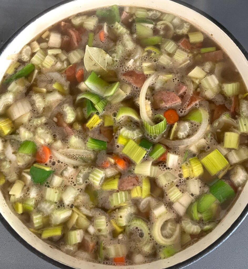 Looking down into a large pot of split pea soup cooking. Soup features cut up carrots, celery, onion, leeks, garlic, and chucks of sausage.