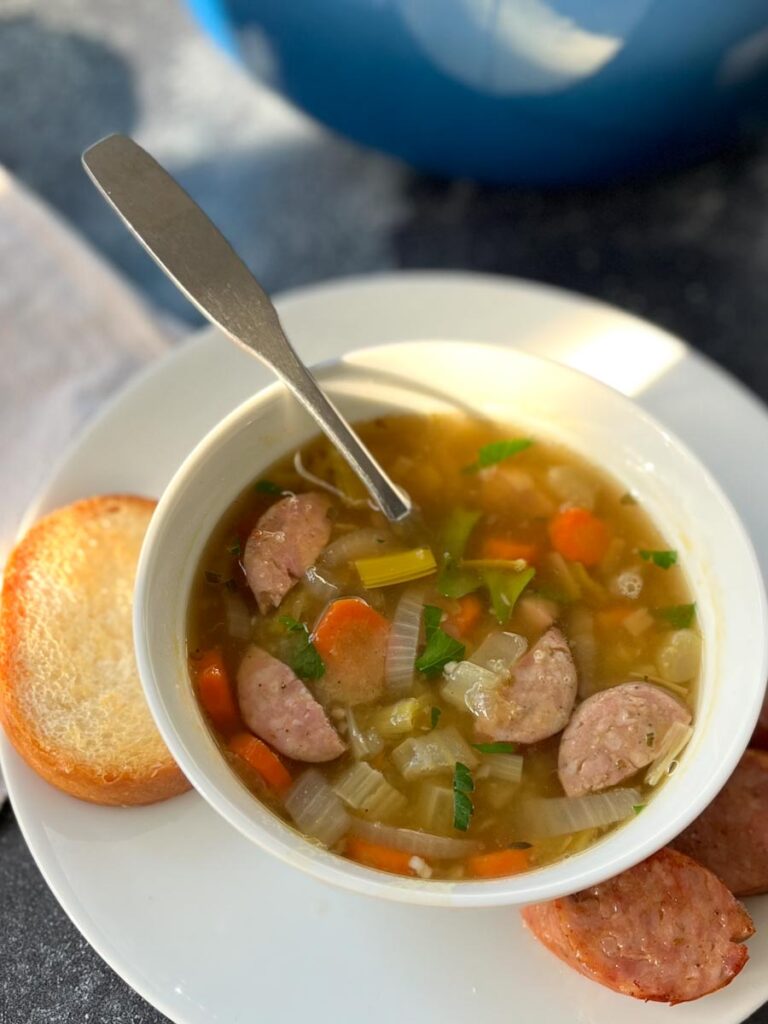 White bowl on a round white plate. Bowl has soup in it consisting of sausage, celery, carrots, and onion. A spoon is resting in the bowl. A piece of bread and 3 slices of sausage are on the plate. There is a light brown cloth napkin next to the plate. Items are on a gray surface.