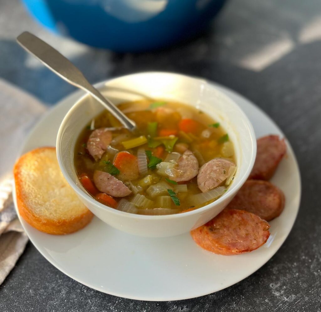 White bowl on a round white plate. Bowl has soup in it consisting of sausage, celery, carrots, and onion. A spoon is resting in the bowl. A piece of bread and 3 slices of sausage are on the plate. There is a light brown cloth napkin next to the plate. Items are on a gray surface.