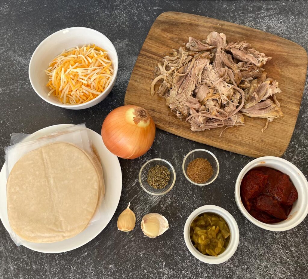 There is a small wooden cutting board with pulled pork on it. There is a white plate with dough discs on it. There are three small white bowls. The largest bowl has shredded yellow and white cheese in it. One bowl has chopped green chiles the third bowl has red chipotles in it. There is a yellow onion and 2 garlic cloves. There are 2 small clear ramekins. One has ground cumin in it, the other has dried oregano in it.
