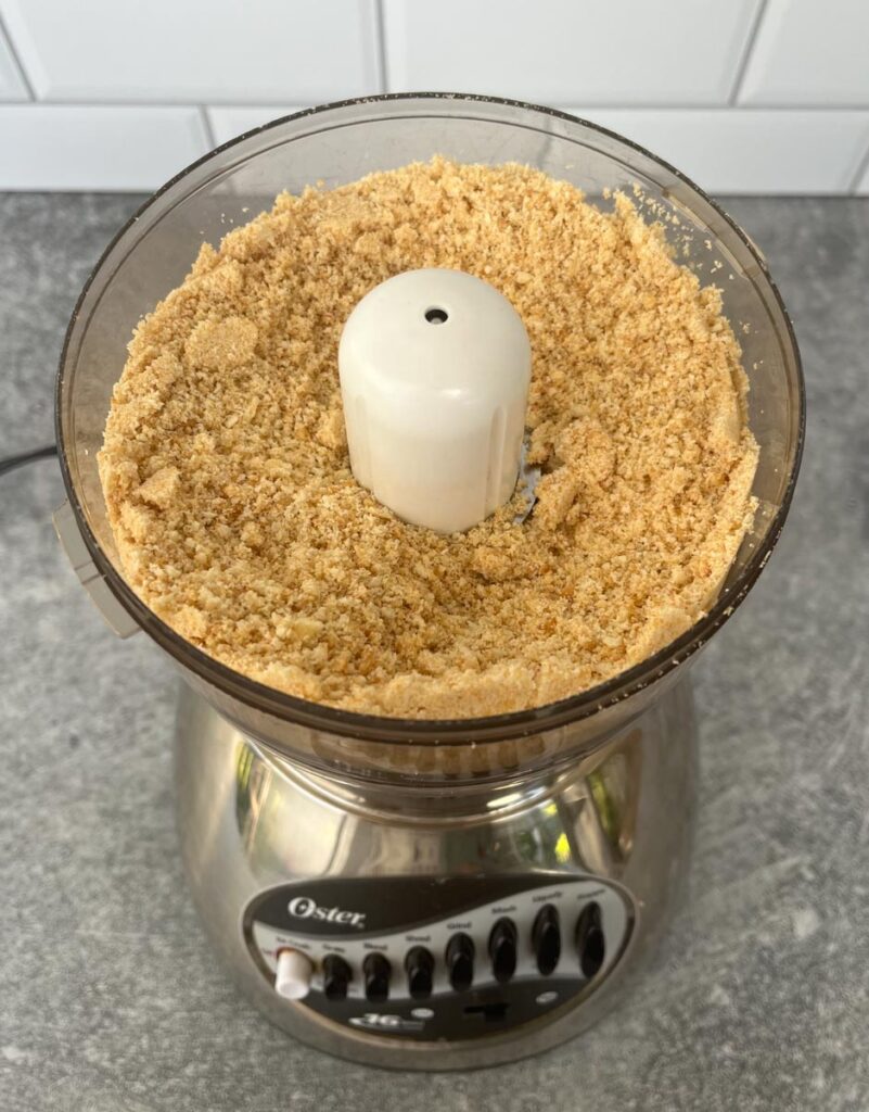 Small food processor with crumbled butter crackers in the bowl section. The processor is on a gray surface with a white subway tile back splash.
