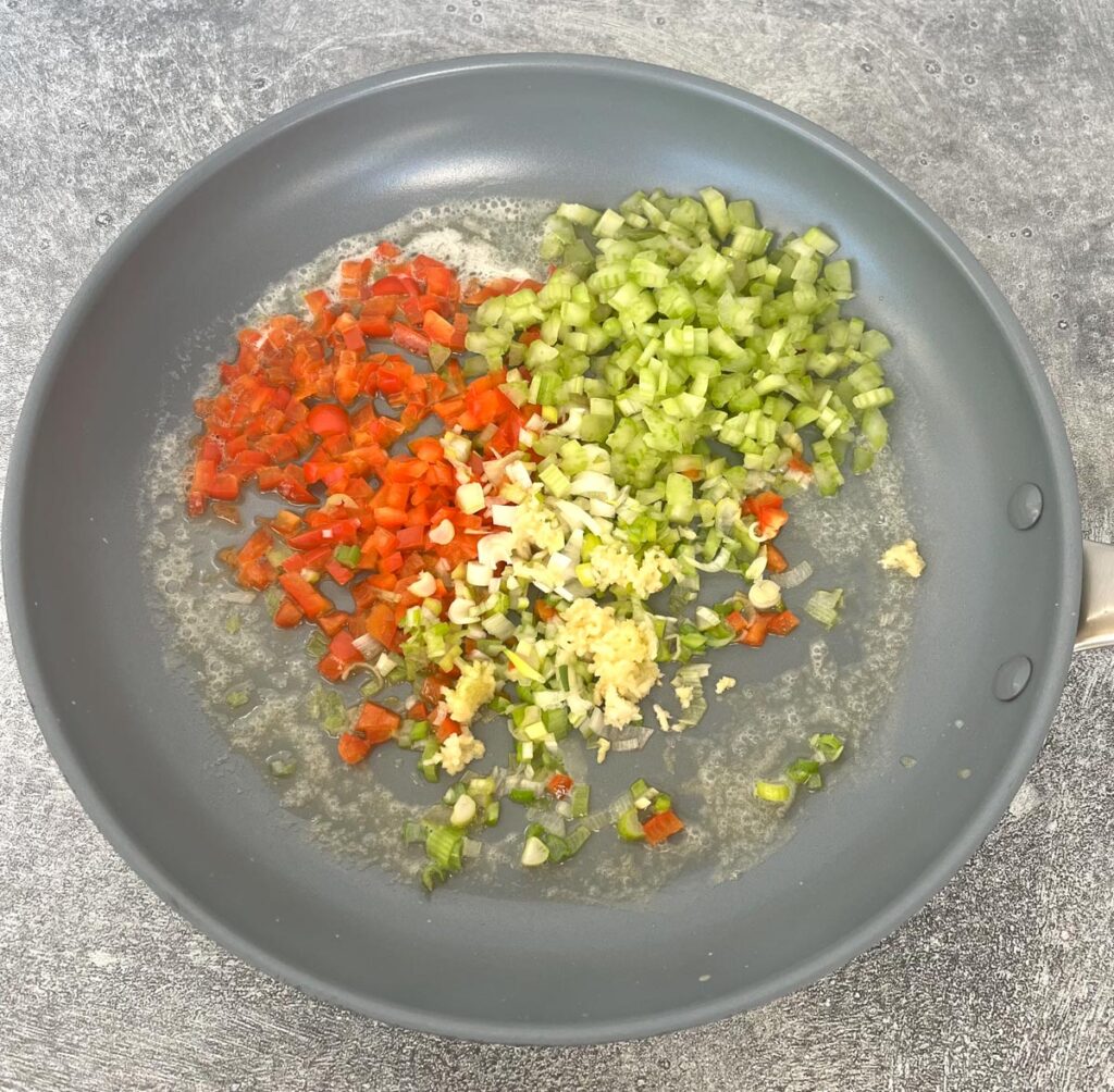 Gray fry pan with chopped celery, chopped red pepper, chopped scallions, and minced garlic. Items are cooking in melted butter. Fry pan is on a gray surface.