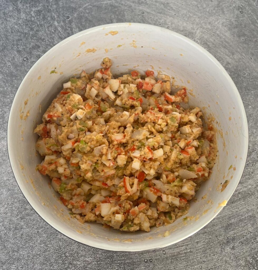 Large white mixing bowl with chopped crabmeat, chopped scallops, cracker crumbs, chopped scallions, chopped celery, chopped red pepper, minced garlic, a raw egg, melted butter, hot sauce, Worcestershire sauce, Old Bay seasoning, and pepper in it. The stuffing is throughly mixed together. The bowl is sitting on a gray surface.