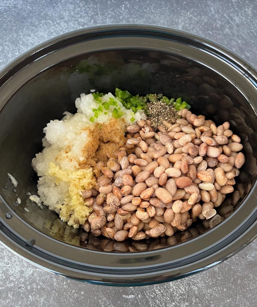 There is a slow cooker with pre soaked pinto beans, chopped white onion, minced garlic, minced jalapeno pepper, ground cumin, and ground pepper in it. The slow cooker is on a gray surface.