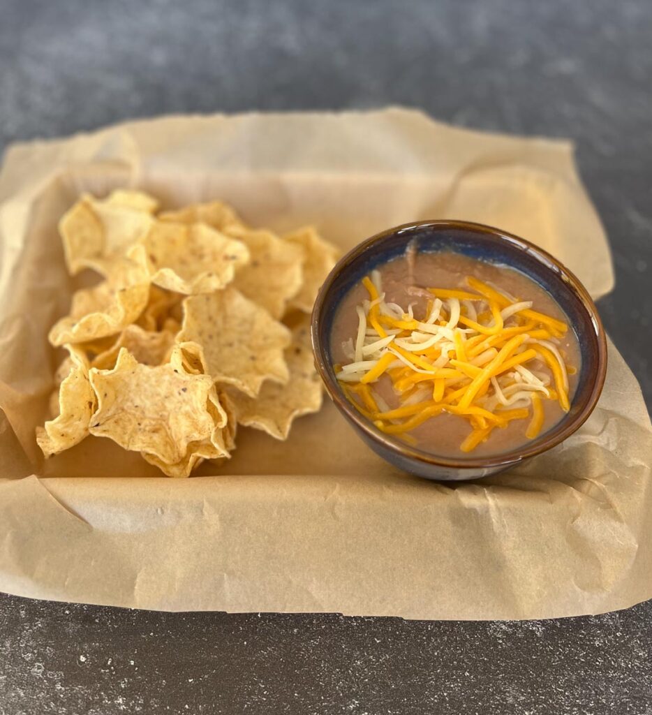 Bowl of charro beans with shredded cheese on top, sitting on a brown paper lined tray. There are Tostitos Scoops Tortilla Chips on the tray next to the bowl. Tray is on a gray surface.