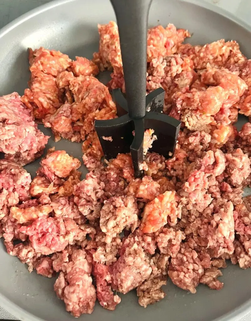 Ground Beef and Sausage In gray fry pan. There is a black ground meat chopper in the pan.