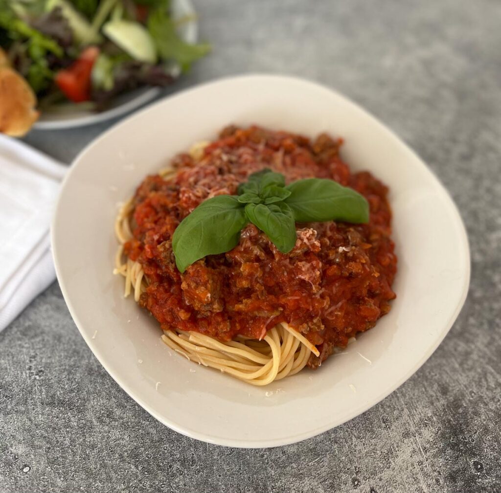 White bowl with spaghetti noodles and tomato and meat sauce on top of noodles. There is fresh basil leaves on top of sauce. In the background there is a white plate with a green salad on it and a white cloth napkin. Items are on a light gray surface.