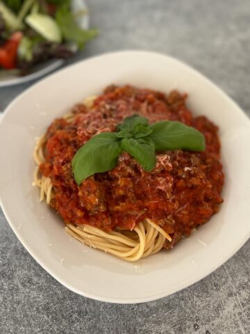 White bowl with spaghetti noodles and tomato and meat sauce on top of noodles. There is fresh basil leaves on top of sauce. In the background there is a white plate with a green salad on it and a white cloth napkin. Items are on a light gray surface.