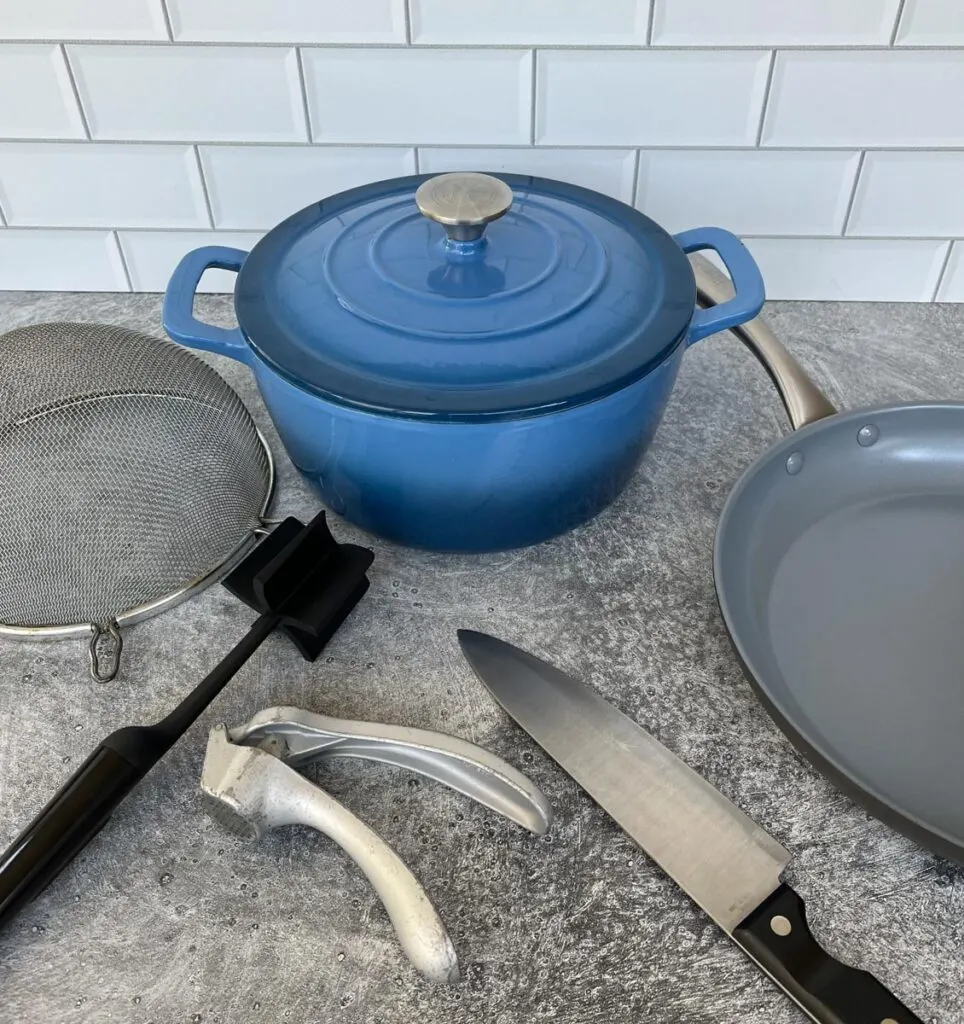 Large blue dutch oven. large gray fry pan, mesh strainer, ground meat chopper, garlic press, and a large kitchen knife. All items are on a light gray surface with a white subway time back splash.